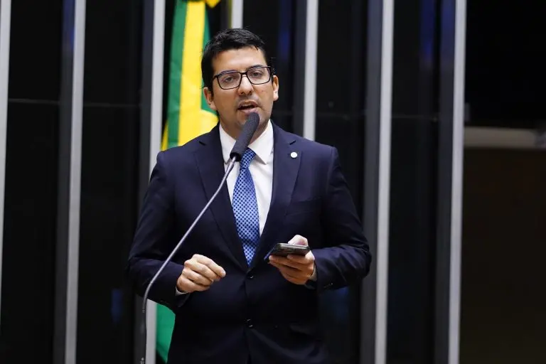 O deputado Marcelo Queiroz discursa ao microfone no plenário da Câmara, vestindo terno azul-marinho e segurando um celular enquanto fala. Ao fundo, as listras verticais do cenário e parte da bandeira do Brasil compõem o ambiente legislativo formal.