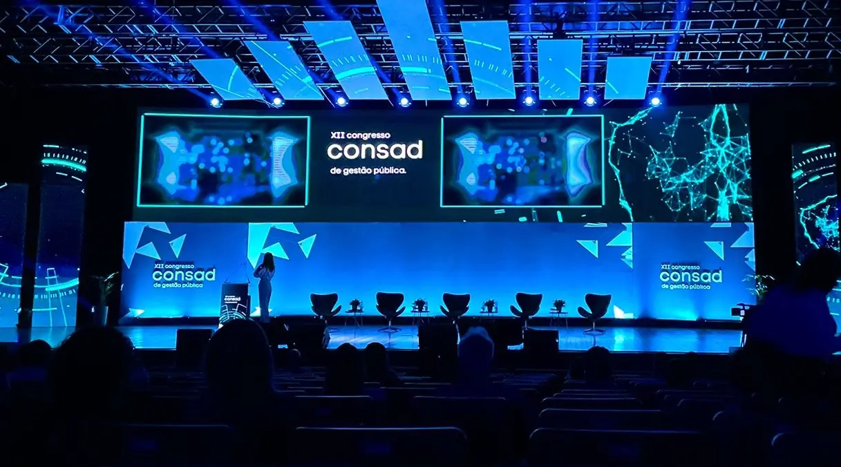 Palco iluminado em azul durante o XII Congresso Consad de Gestão Pública, com telões, cadeiras para debate e público sentado na plateia.