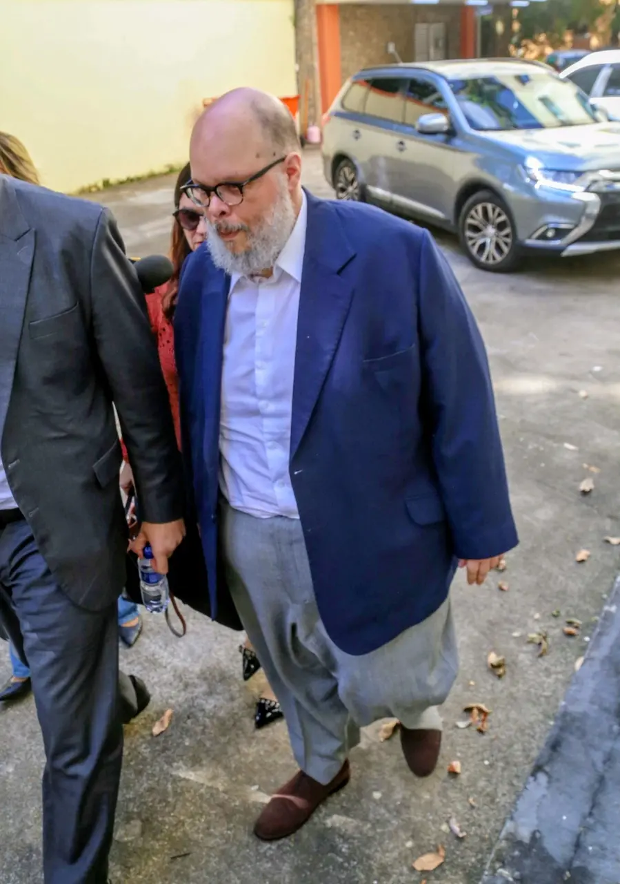 Pessoa de terno azul e camisa clara caminha em área externa pavimentada, acompanhada por outras pessoas, enquanto um microfone se aproxima lateralmente; ao fundo, veículo estacionado e fachada de prédio.