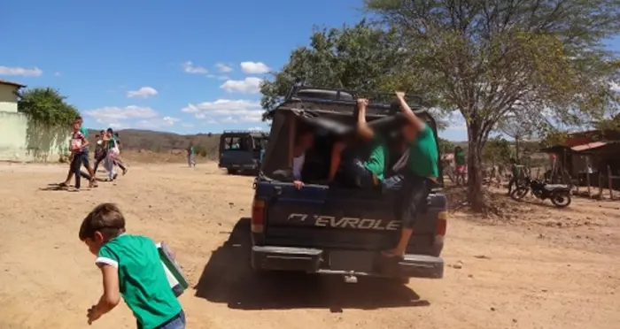 Grupo de estudantes com uniformes verdes sendo transportados na caçamba aberta de uma caminhonete Chevrolet antiga em uma estrada de terra. Algumas crianças aparecem penduradas na estrutura externa do veículo sob sol forte, enquanto outras caminham ao fundo em uma área rural árida com vegetação esparsa. As faces dos jovens na imagem estão borradas para preservar a identidade.