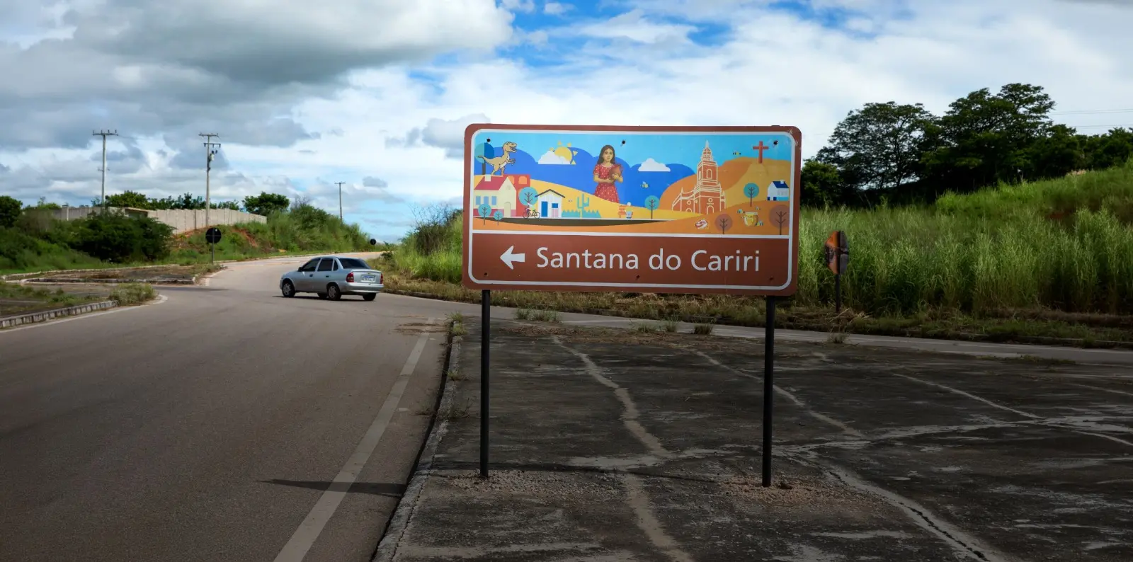 Na imagem, fotografia em plano médio de uma placa de sinalização turística à beira de uma estrada, indicando a direção para Santana do Cariri. A placa é retangular com fundo marrom e possui uma ilustração colorida no topo que retrata elementos da região: um dinossauro, o sol, uma mulher com um pote de barro, uma igreja e uma cruz. Abaixo da ilustração, há uma seta branca apontando para a esquerda seguida pelo nome 
