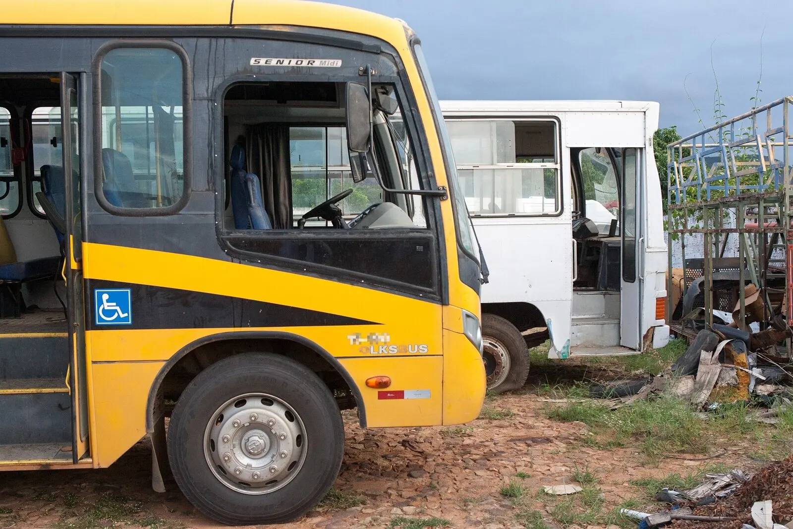 Dois ônibus escolares em estado de abandono ou manutenção estacionados em um terreno de terra e pedregulhos. O primeiro é amarelo com detalhes pretos e símbolo de acessibilidade, sem o vidro da janela do motorista, enquanto o segundo, ao fundo, é totalmente branco e parece estar sendo desmontado em meio a sucatas metálicas.