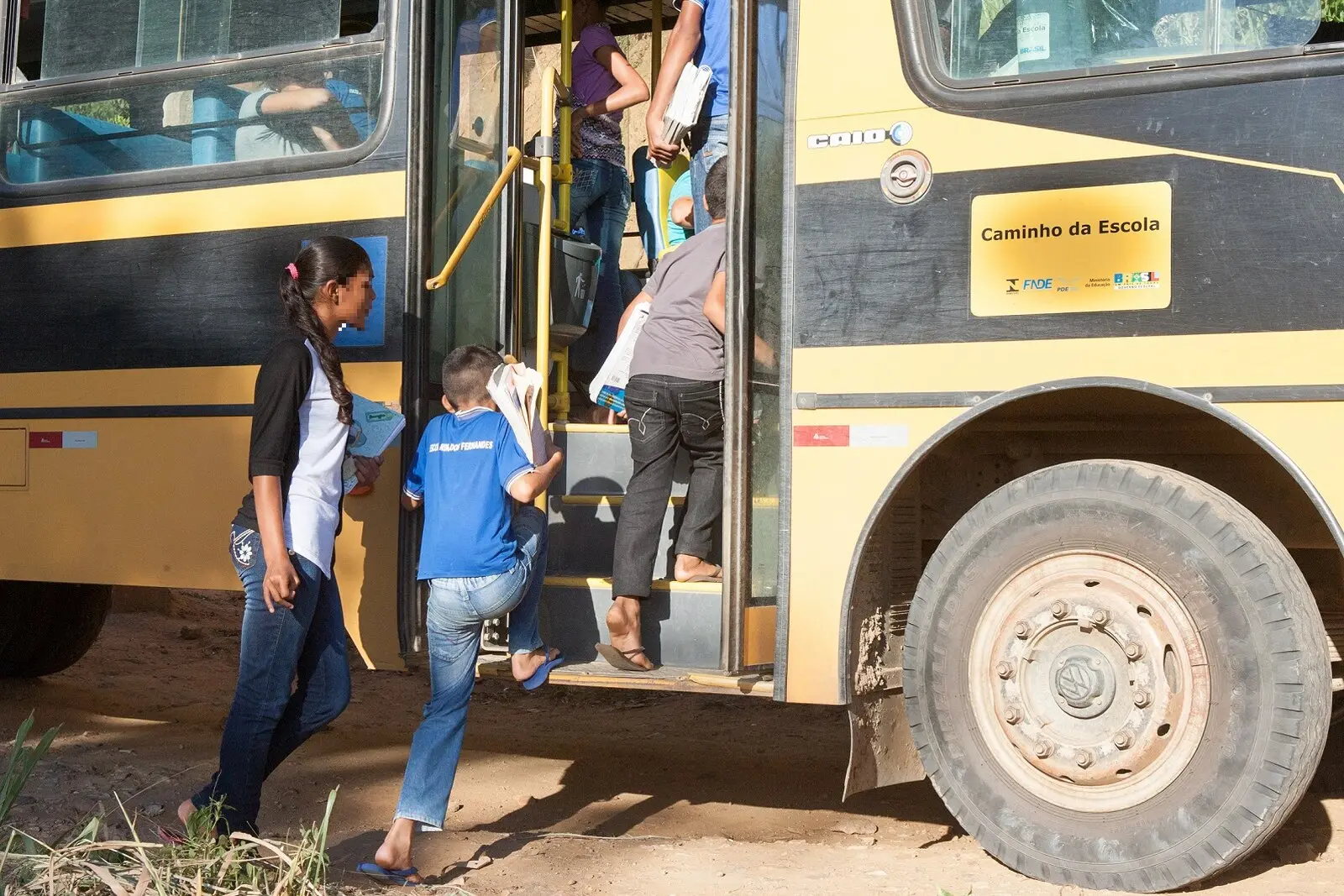 Crianças e adolescentes carregando cadernos e livros sobem os degraus de um ônibus escolar amarelo e preto com o selo Caminho da Escola. A cena ocorre em uma estrada de terra batida sob luz solar intensa, destacando um dos pneus dianteiros sujo de poeira e os estudantes vestindo roupas casuais.