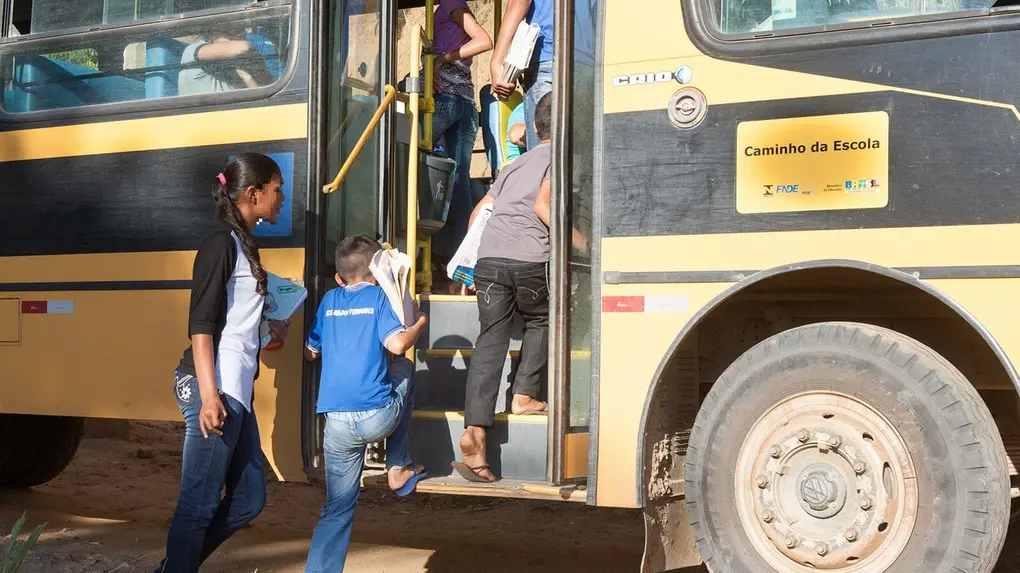 Crianças e adolescentes carregando cadernos e livros sobem os degraus de um ônibus escolar amarelo e preto com o selo Caminho da Escola. A cena ocorre em uma estrada de terra batida sob luz solar intensa, destacando um dos pneus dianteiros sujo de poeira e os estudantes vestindo roupas casuais.