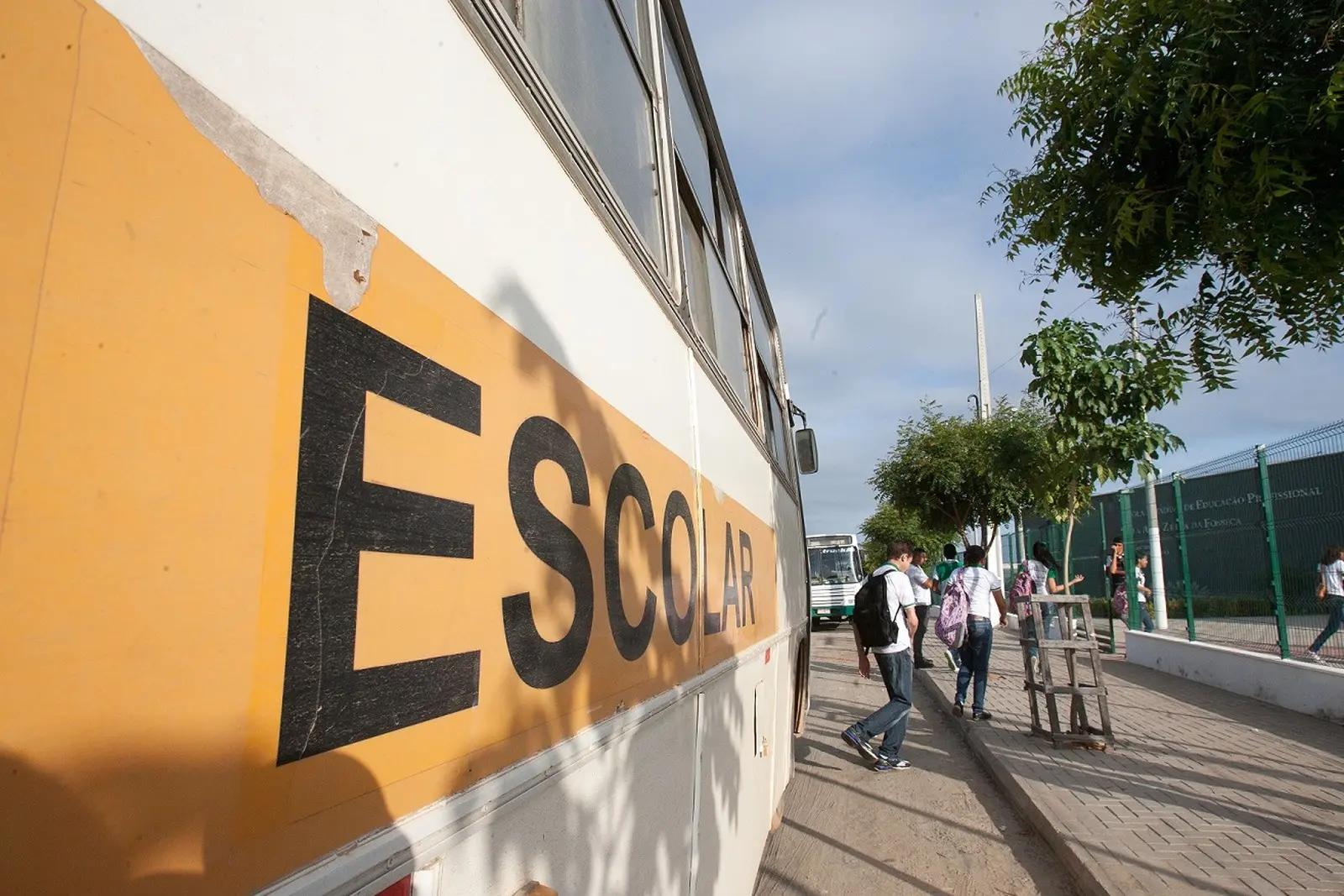 Vista lateral em perspectiva de um ônibus branco com uma grande faixa amarela onde se lê ESCOLAR em letras pretas. Na calçada ao lado, estudantes de uniforme branco e mochilas caminham em direção a uma escola protegida por grades verdes, sob um céu claro com poucas nuvens.