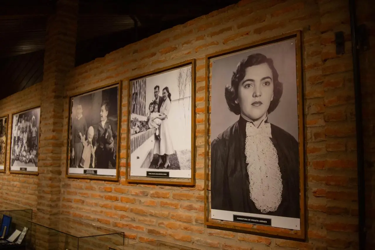 Visitantes observam fotografias e objetos expostos em espaço museológico de paredes de tijolo aparente e iluminação amarela. Entre eles, um homem segura uma criança no colo enquanto acompanha a exposição.