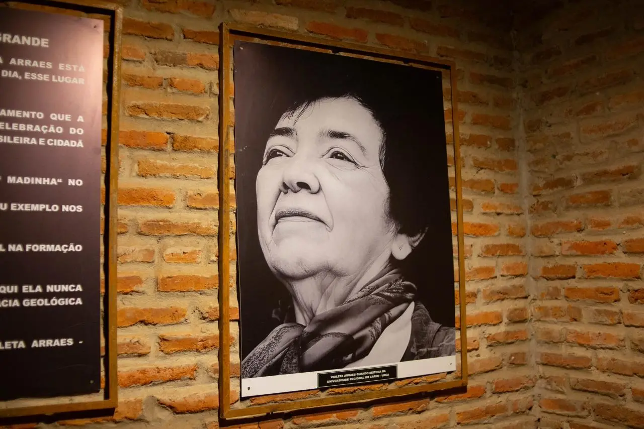 Retrato em preto e branco de uma mulher idosa exposto em parede de tijolo aparente em espaço cultural. Ao lado da fotografia, há um painel com texto informativo sobre a homenageada.