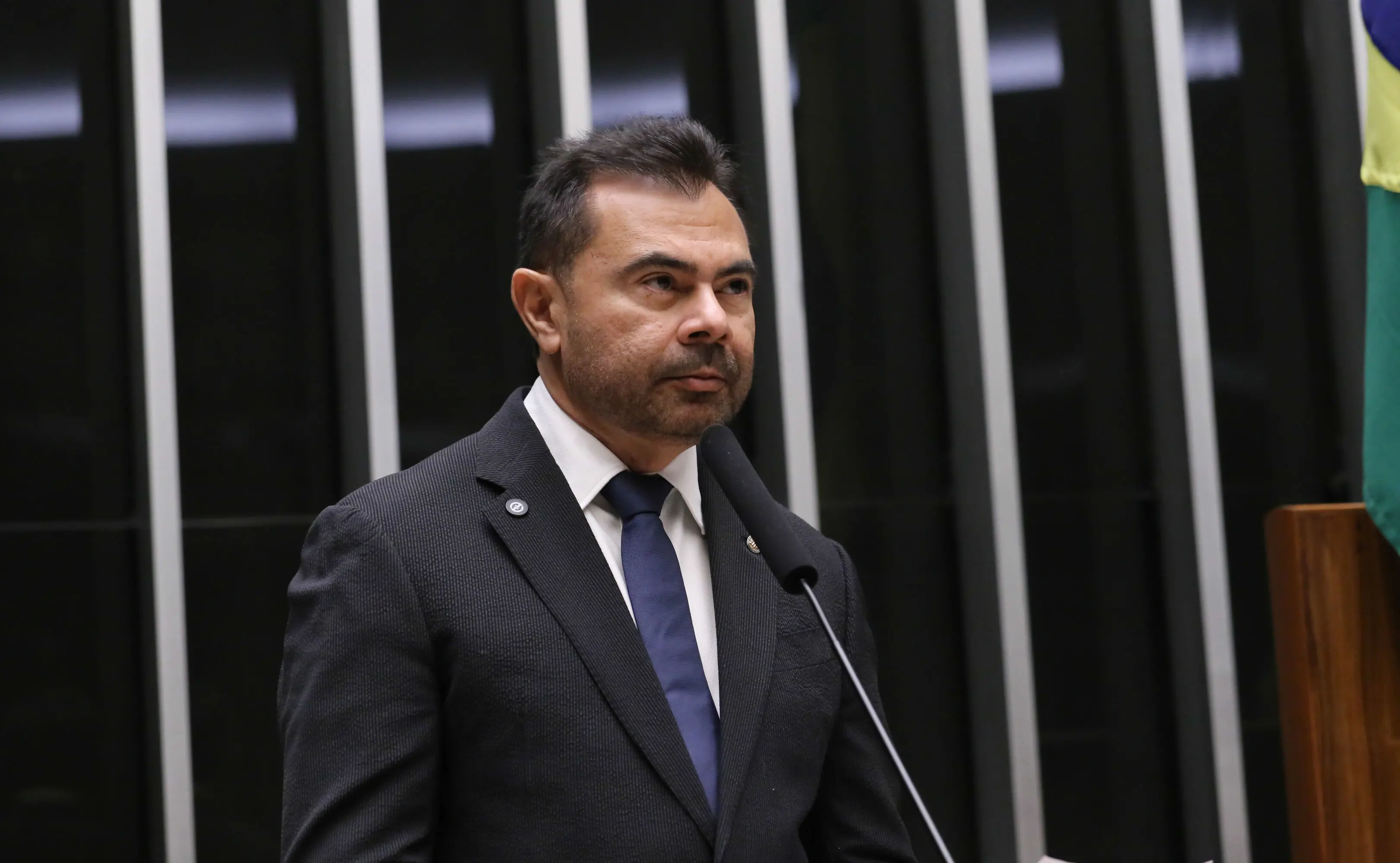 Homem de terno escuro e gravata azul fala ao microfone em tribuna da Câmara dos Deputados. Ao fundo, aparecem painéis verticais escuros e parte de uma bandeira do Brasil.