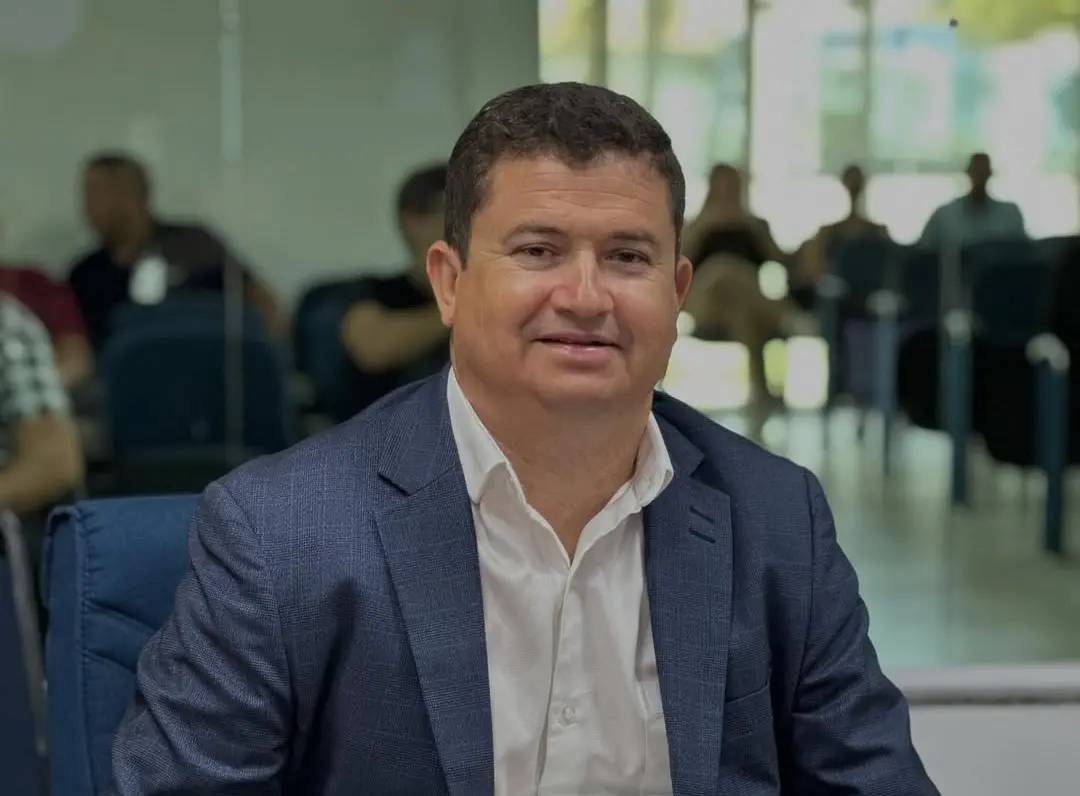 Homem de cabelos curtos escuros, usando blazer azul-marinho e camisa branca, sentado em uma cadeira em ambiente interno. Ele olha para a câmera com expressão leve e sorridente. Ao fundo, há pessoas desfocadas sentadas em cadeiras, em um espaço com paredes de vidro e iluminação natural.