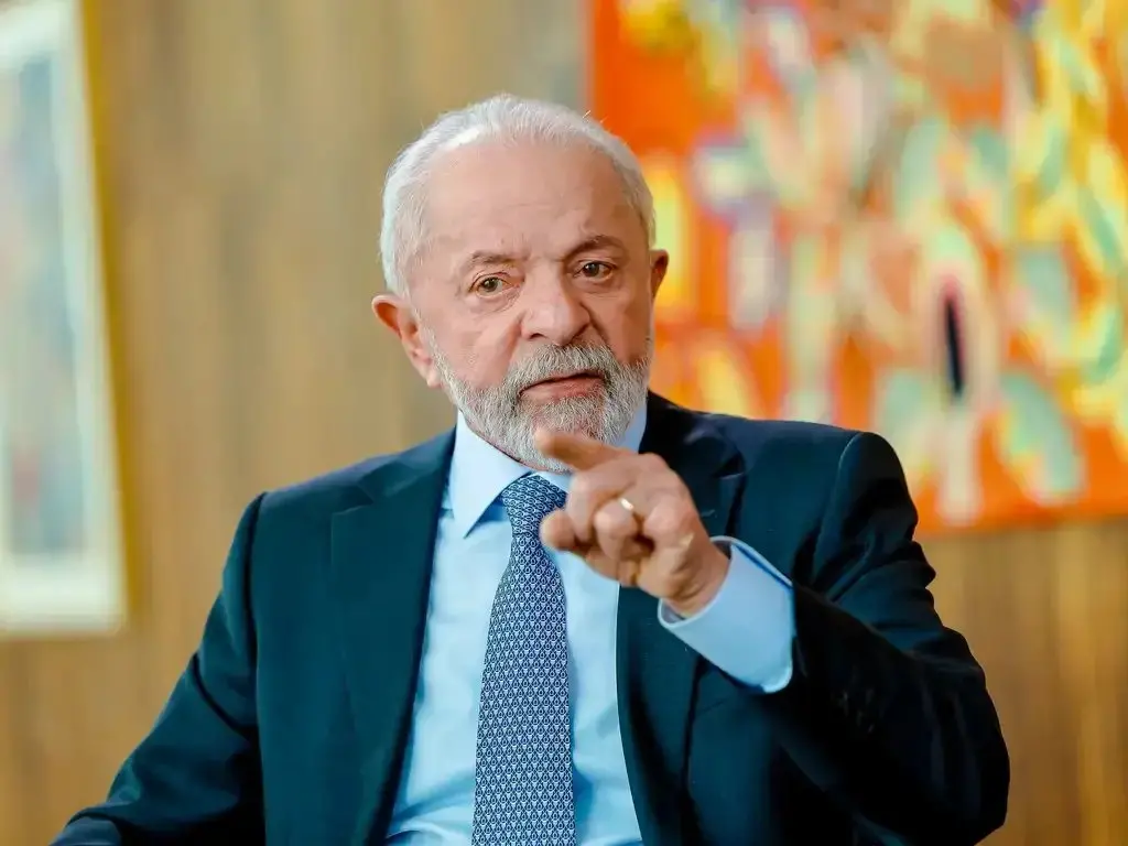 foto do presidente Lula vestindo terno azul, gesticulando com o dedo durante conversa, em ambiente com decoração colorida ao fundo.