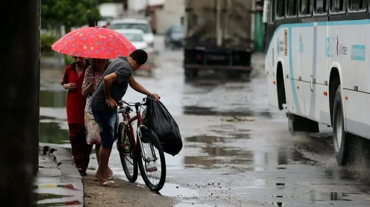 Na imagem, captura em um dia de chuva sob a luz natural difusa. No centro, um homem vestindo camiseta cinza e bermuda jeans manuseia uma bicicleta vermelha próxima ao meio-fio. Atrás dele, um homem com camisa vermelha e uma mulher com blusa estampada dividem um guarda-chuva vermelho vibrante. A rua de asfalto está molhada, refletindo os veículos e o céu cinzento. À direita, a lateral de um ônibus urbano branco em movimento ocupa parte da composição, com gotas de água sendo dispersadas pelos pneus. O fundo mostra outros carros e árvores de forma desfocada.
