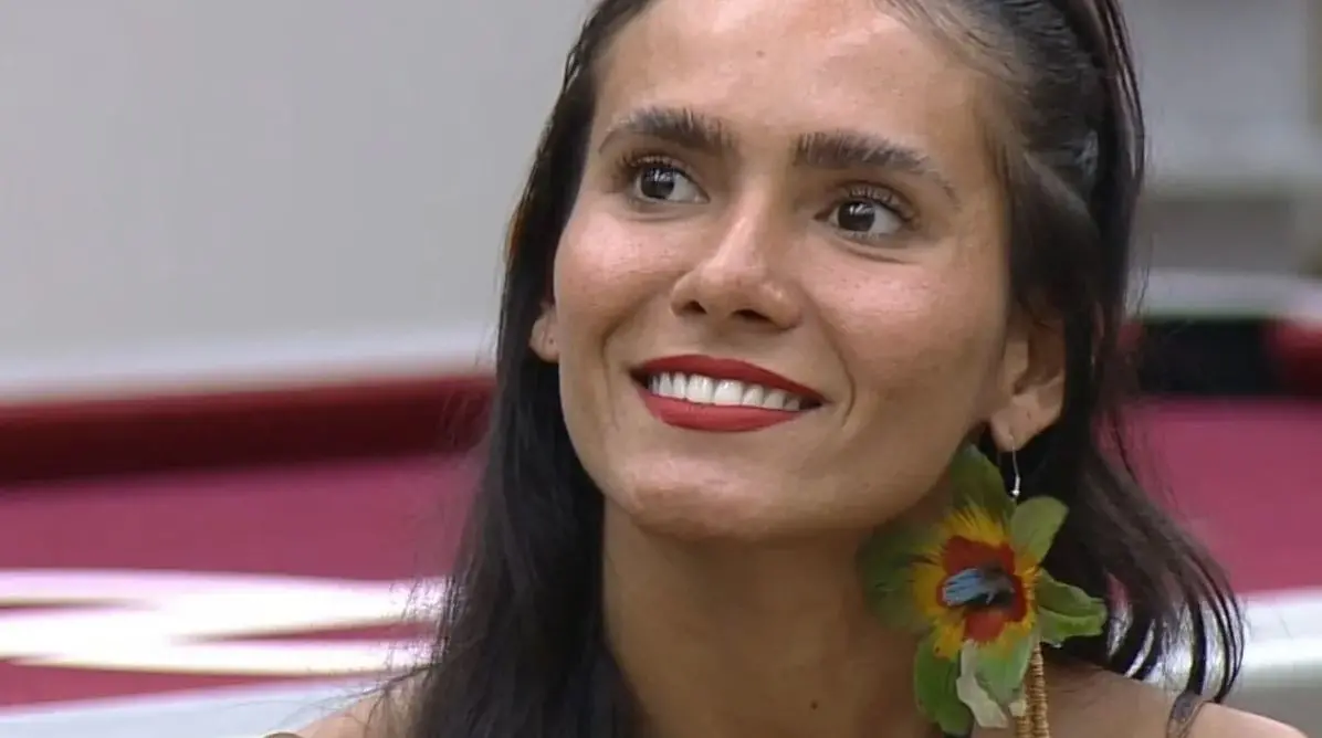 Close em participante usando brincos grandes em formato de flor, com cabelo preso para trás, lábios com batom vermelho e dentes aparentes. Fundo desfocado em tons claros e rosados.
