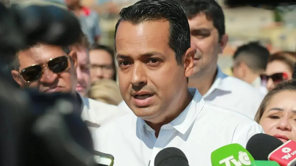 Homem de camisa branca fala com a imprensa ao ar livre, cercado por repórteres e microfones de emissoras de TV. Ele aparece em primeiro plano, olhando para frente durante uma entrevista coletiva. Ao fundo, há várias pessoas acompanhando a fala, algumas usando óculos escuros. A imagem tem foco no rosto do entrevistado e nos microfones posicionados à frente dele.