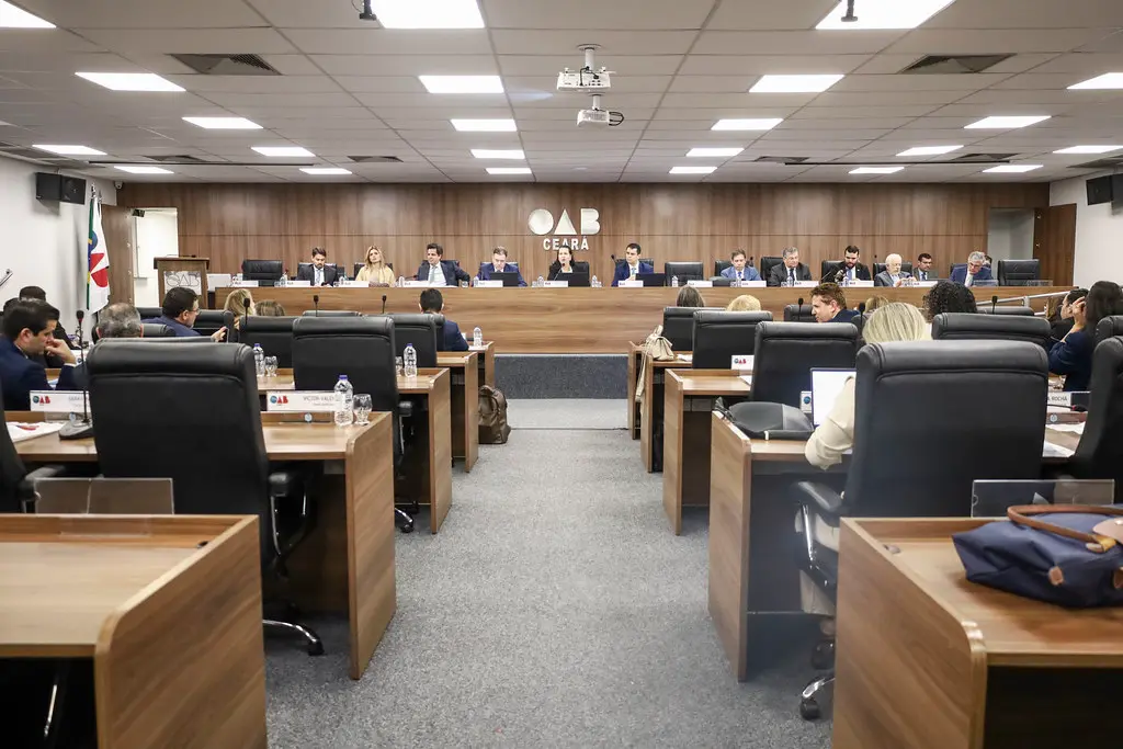 Auditório da OAB Ceará durante sessão plenária, com advogados e conselheiros sentados em mesas e cadeiras organizadas em fileiras. Ao fundo, uma bancada elevada reúne integrantes da mesa diretora sob o logotipo da OAB Ceará na parede de madeira. O ambiente é amplo, iluminado e possui projetor no teto.