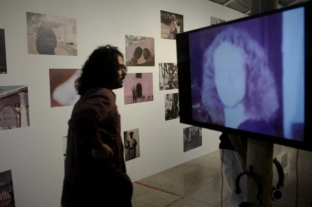 Um visitante observa a exposição sobre Jomard Muniz de Britto no CCBNB, caminhando em frente a uma parede branca repleta de fotografias fixadas que retratam cenas diversas. Em primeiro plano, uma tela de vídeo exibe um rosto em tons de roxo, complementando a experiência imersiva da mostra.