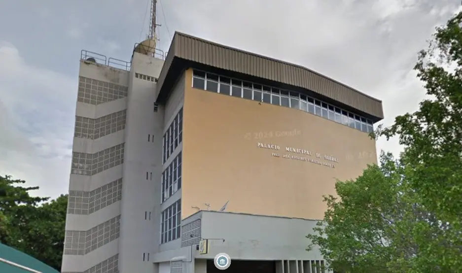 Fachada do Palácio Municipal de Sobral, prédio administrativo de vários andares com estrutura em tons de bege e branco. O edifício possui janelas amplas, torre lateral com antena no topo e árvores ao redor. No centro da fachada, há a identificação “Palácio Municipal de Sobral Prefeito José Euclides Ferreira Gomes”.