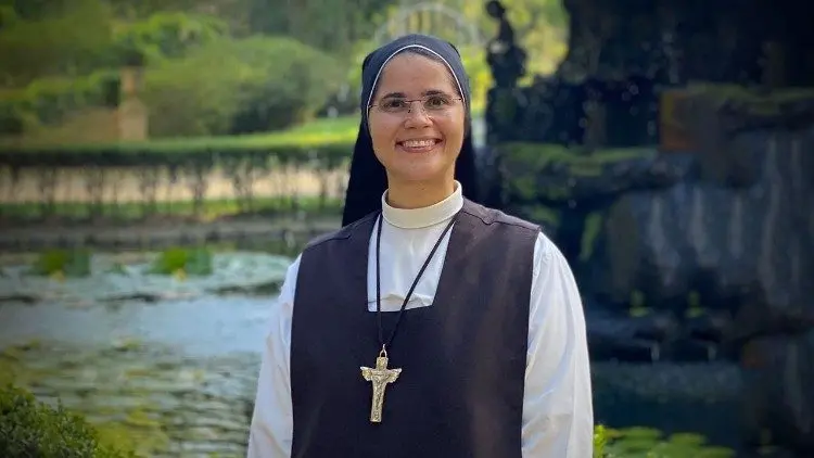 Freira usando hábito religioso preto e branco, com véu e cruz de madeira no peito, sorrindo ao ar livre em frente a um jardim e um lago ao fundo.