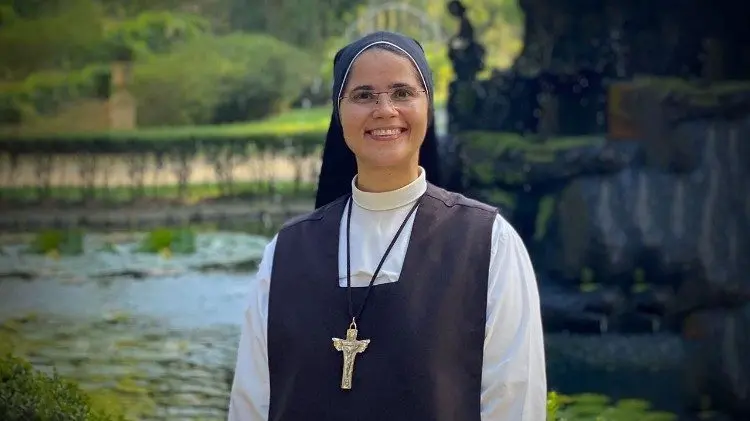 Freira usando hábito religioso preto e branco, com véu e cruz de madeira no peito, sorrindo ao ar livre em frente a um jardim e um lago ao fundo.