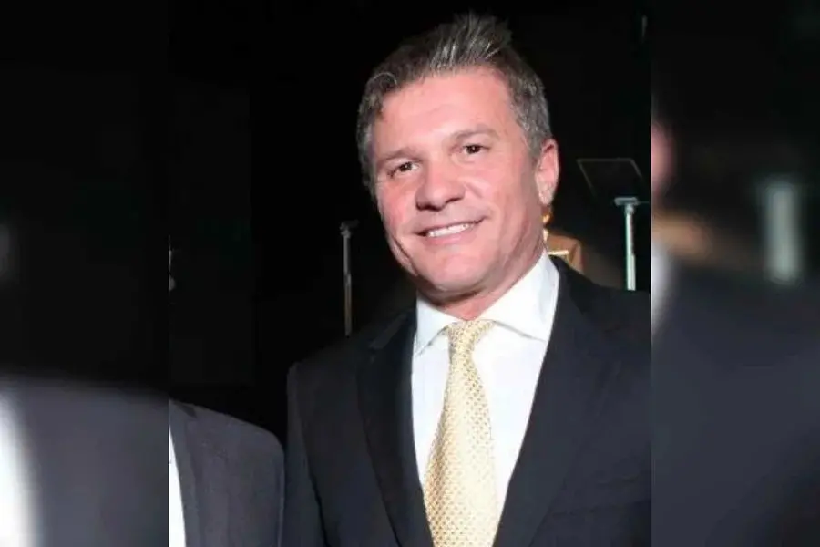 Homem adulto em evento noturno, usando terno escuro, camisa clara e gravata bege, posando para foto. Ele tem cabelo grisalho e expressão sorridente.