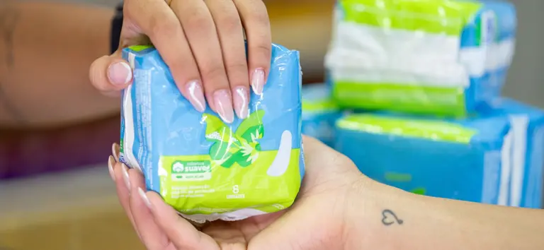 Pessoa segurando um pacote de lenços de papel azul com detalhes verdes, enquanto outras embalagens do mesmo tipo ficam ao fundo na mesa, em foto com foco nas mãos.