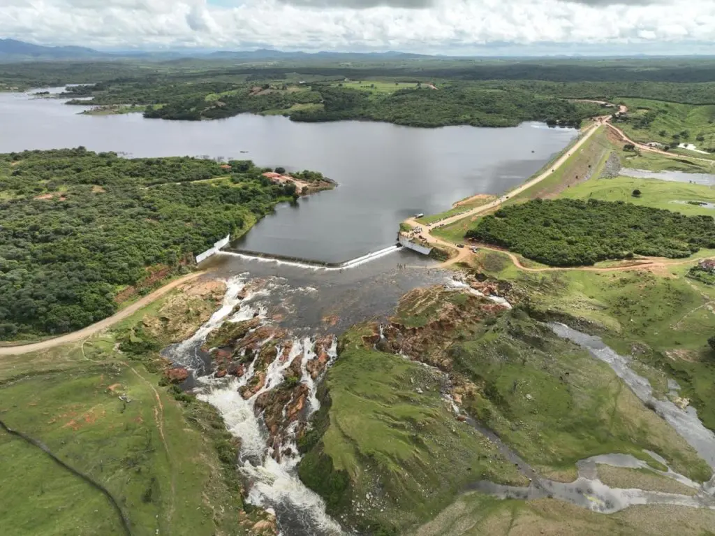 Foto aérea do açude Serafim Dias em Mombaça vertendo água sobre o sangradouro, cercado por vegetação densa e estradas de terra sob céu claro.