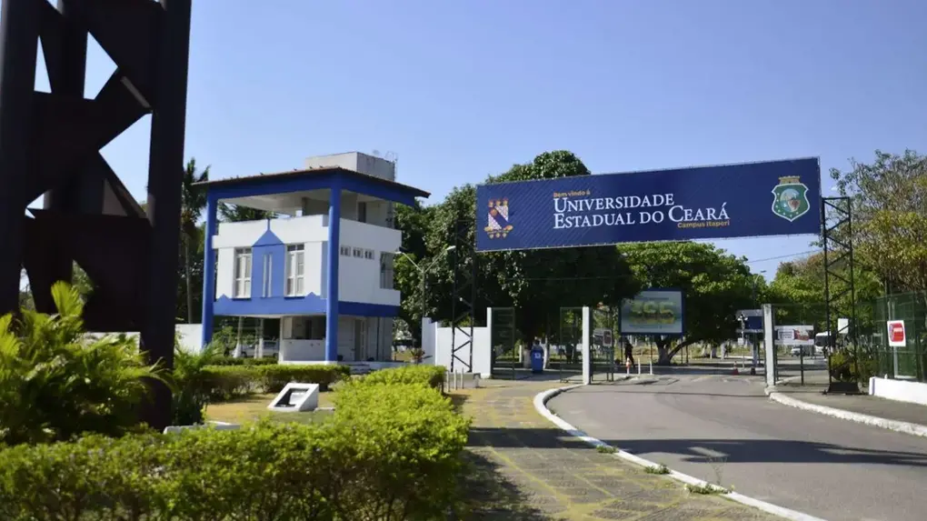 Entrada da Universidade Estadual do Ceará (UECE), campus Itaperi, com pórtico de identificação azul à direita, trazendo o nome da instituição. À esquerda, há uma guarita branca e azul de dois pavimentos. A via de acesso é asfaltada, com calçada, canteiros com arbustos e árvores ao redor, sob céu claro e ensolarado.