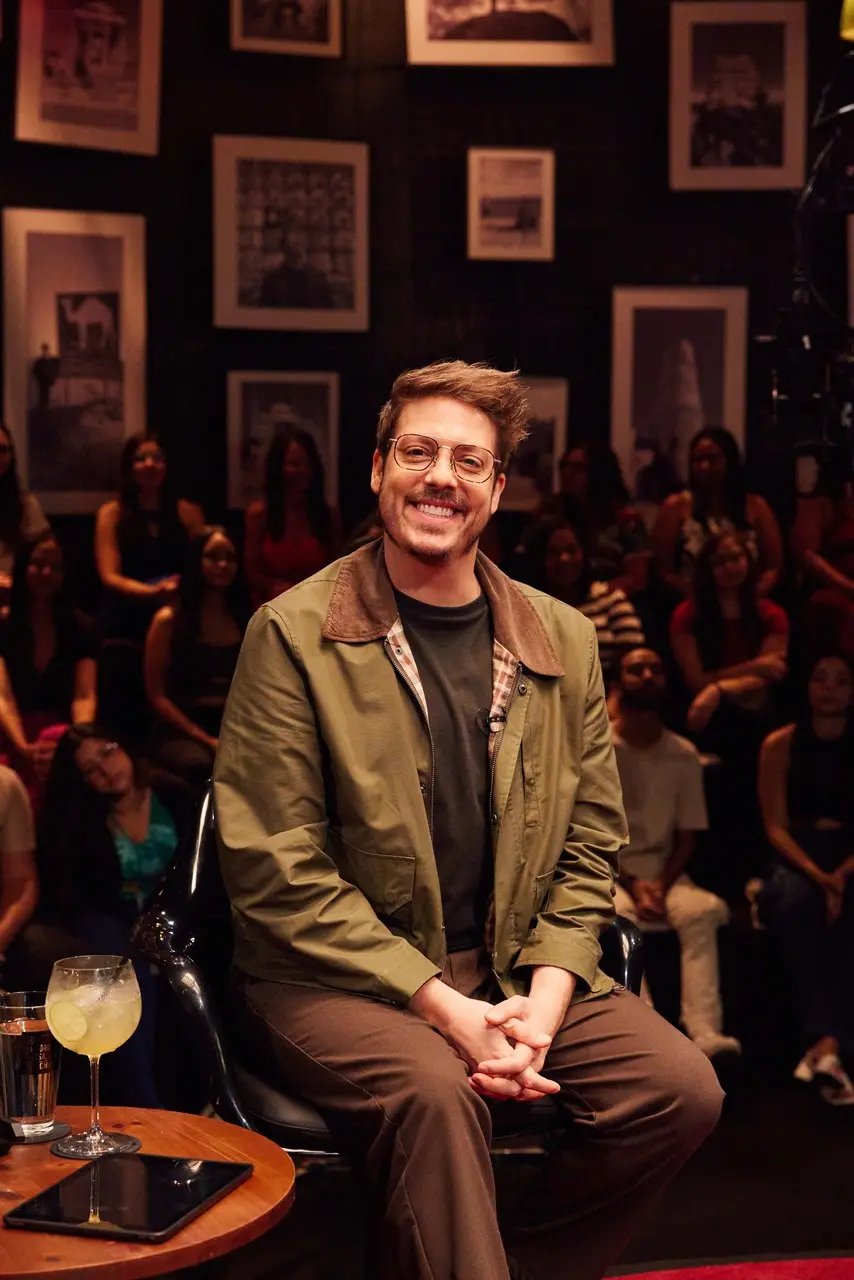 O apresentador Fábio Porchat aparece sorridente, sentado em uma cadeira preta no centro do estúdio do programa 'Que História É Essa, Porchat?'. Ele veste uma jaqueta verde-oliva com gola marrom sobre uma camiseta escura e calça marrom, tendo ao fundo uma parede escura decorada com quadros emoldurados e a plateia desfocada.