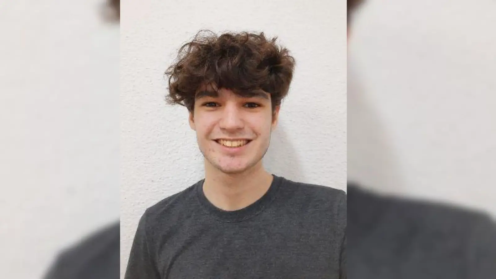 Jovem sorrindo para a câmera, com cabelos castanhos cacheados e camiseta cinza escura, em frente a uma parede branca.