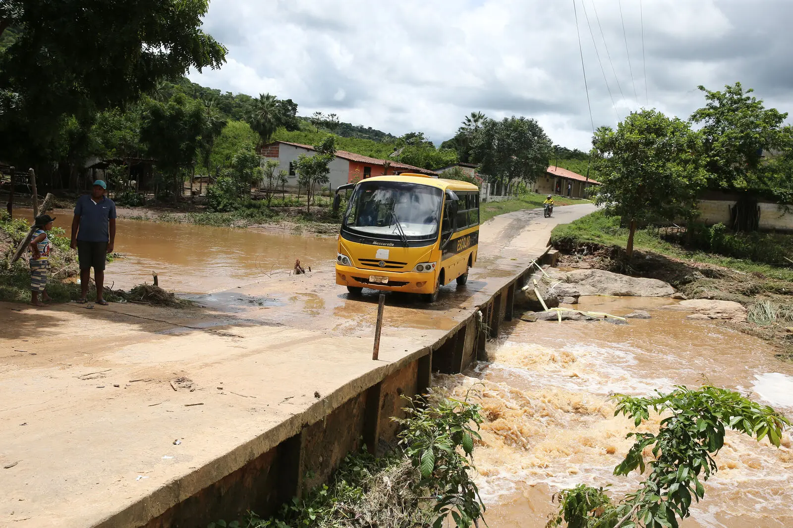 Pequeno ônibus escolar amarelo atravessa ponte rústica sobre riacho barrento e caudaloso. Na margem de terra, um homem e um menino observam a cena, com um vilarejo rural e vegetação densa ao fundo sob céu nublado.