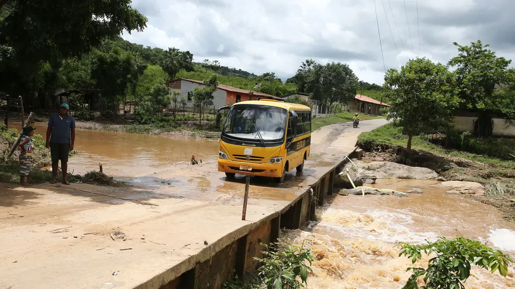 Pequeno ônibus escolar amarelo atravessa ponte rústica sobre riacho barrento e caudaloso. Na margem de terra, um homem e um menino observam a cena, com um vilarejo rural e vegetação densa ao fundo sob céu nublado.