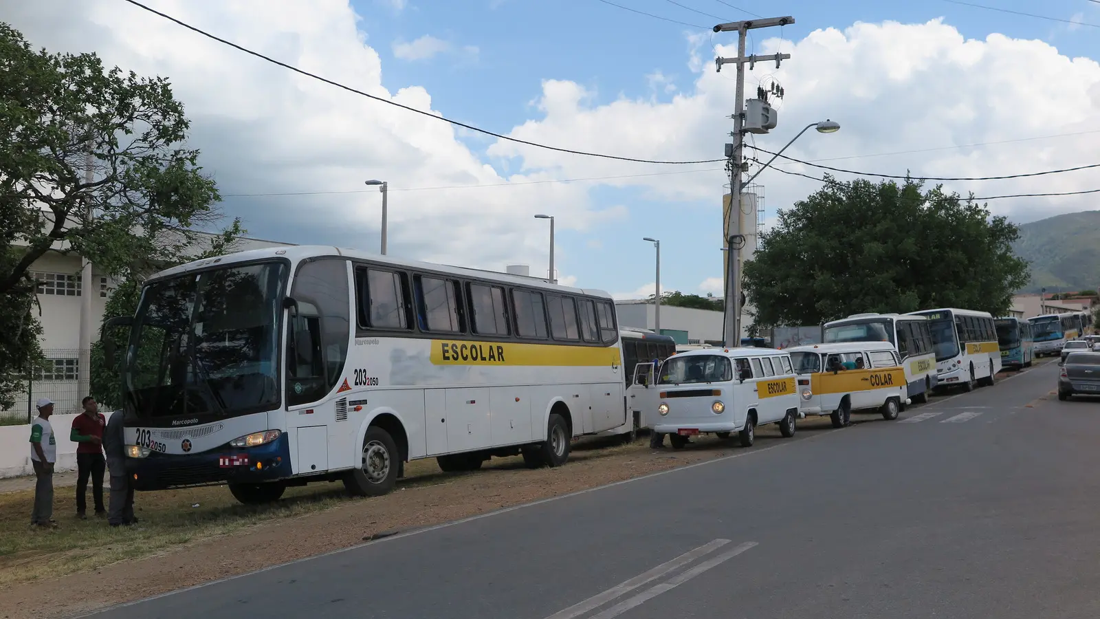 Fila de veículos de transporte escolar, incluindo um ônibus grande e vans Kombi brancas e amarelas com a faixa ESCOLAR, estacionados para vistoria na margem de uma estrada. Três homens conversam perto da frente do primeiro ônibus sob céu nublado.