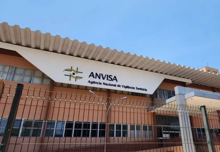 A fachada da sede da Anvisa apresenta o logotipo e o nome da Agência Nacional de Vigilância Sanitária em destaque em um painel branco inclinado sob um telhado metálico ondulado. O edifício de tijolos aparentes e janelas de vidro é visto por trás de uma grade metálica de proteção, sob um céu azul limpo e iluminação solar direta.