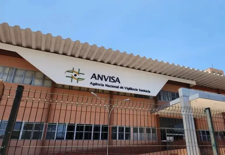 A fachada da sede da Anvisa apresenta o logotipo e o nome em destaque em um painel branco inclinado sob um telhado metálico ondulado. O edifício de tijolos aparentes e janelas de vidro é visto por trás de uma grade metálica de proteção, sob um céu azul limpo e iluminação solar direta. Imagem para matéria onde Unilever informou Agência sobre irregularidade da Ypê.