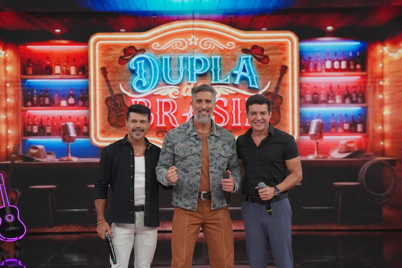 A dupla Marcos e Belutti e o apresentador Marcos Mion (ao centro) sorriem e seguram microfones no centro de um cenário temático que remete a um bar rústico, com um grande letreiro luminoso escrito 'Dupla do Brasil' ao fundo. A composição destaca elementos sertanejos, como garrafas em prateleiras iluminadas, chapéus de cowboy e violões, criando uma atmosfera vibrante e festiva.