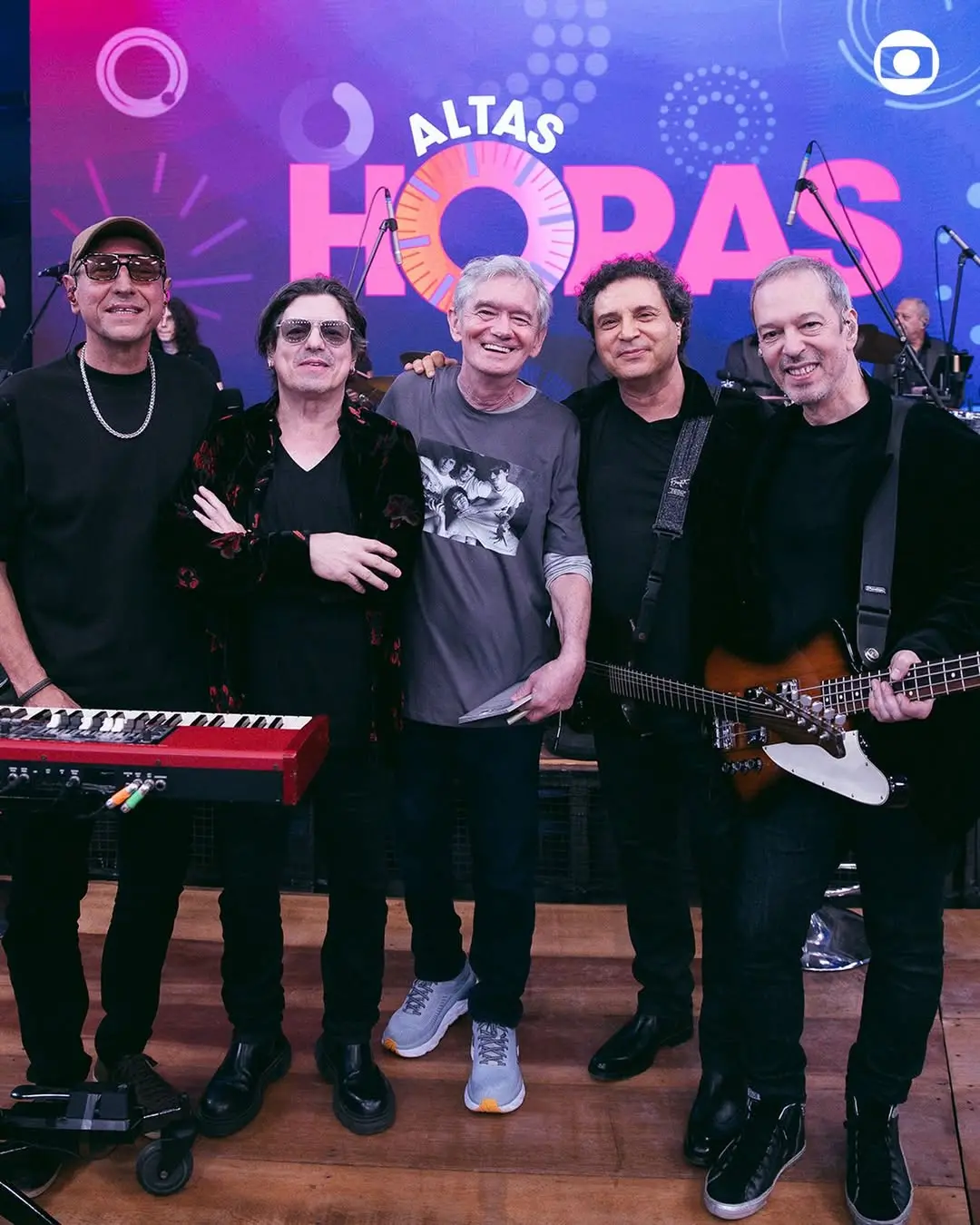 Serginho Groisman e os músicos do Barão Vermelho posam sorridentes no palco do 'Altas Horas', com o logotipo colorido do programa brilhando ao fundo. A imagem destaca a união do grupo entre instrumentos como teclado e baixo, criando uma atmosfera de celebração musical no cenário da TV Globo.