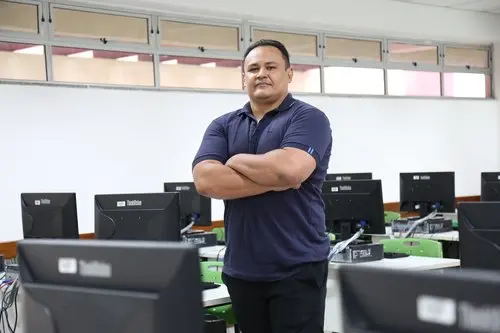 Homem de camisa azul com braços cruzados em laboratório de informática, ao lado de computadores com monitores e teclados, em ambiente de escritório ou sala técnica.