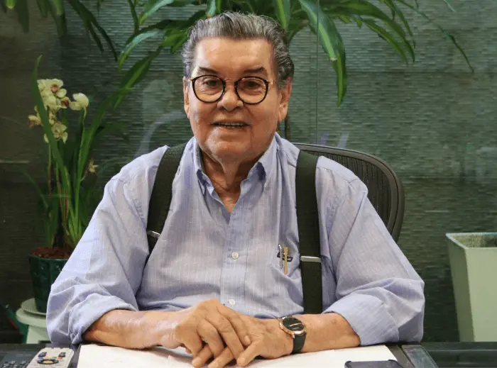 O empresário Francisco 'Dico' de Araújo Carneiro sorri sentado à sua mesa de trabalho, vestindo uma camisa social clara com suspensórios escuros e óculos de armação redonda. Ao fundo, plantas decorativas e uma parede de textura cinza compõem o ambiente de escritório sóbrio e profissional.