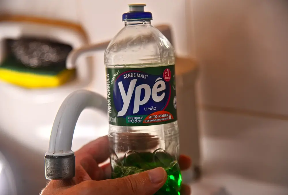 Frasco de detergente transparente com líquido verde, rotulado “Ypê Limão”, sendo segurado por uma mão ao lado de uma torneira de pia.