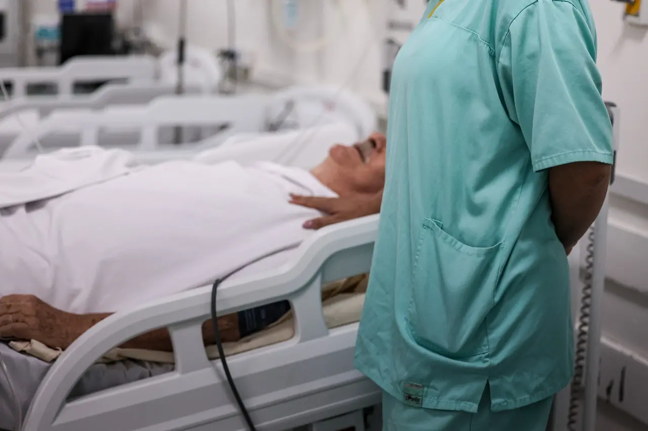 Mulher, profissional da área da saúde, com a mão no ombro esquerdo de um homem idoso, de bigode grisalho, deitado em uma maca hospitalar.