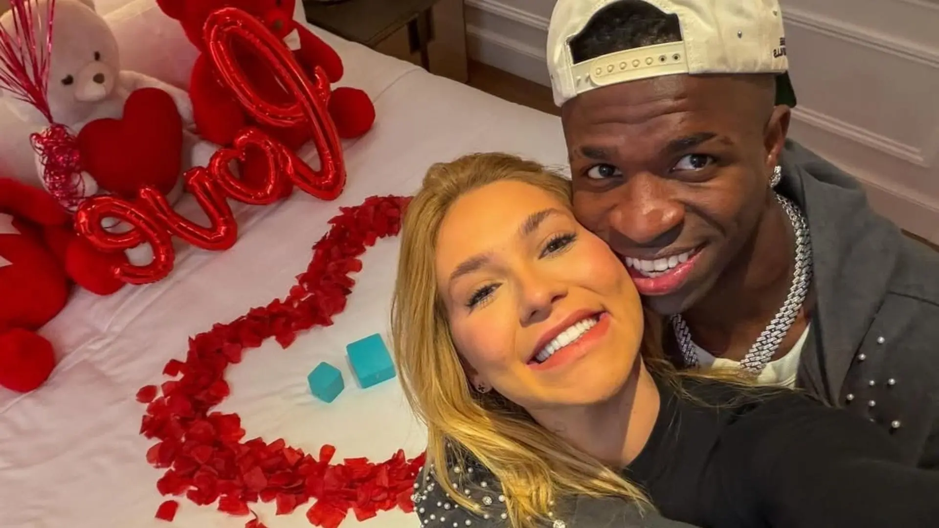 Casal tira selfie sorrindo em quarto decorado com tema romântico, com pétalas de rosas formando um coração sobre a cama, balões vermelhos em formato de letras ao fundo e bichos de pelúcia.