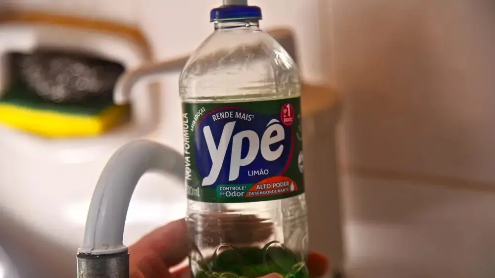 Uma mão segura um frasco de detergente Ypê Limão em destaque, posicionado em frente a uma torneira de cozinha com fundo levemente desfocado. O rótulo verde e azul do produto evidencia a marca e sua finalidade, compondo uma cena cotidiana de limpeza doméstica.