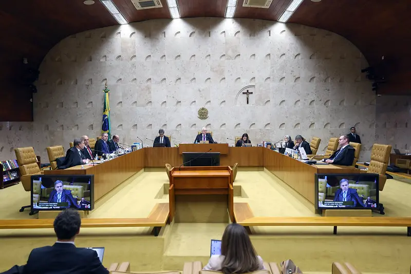 Plenário do STF durante sessão de julgamento, com ministros sentados em bancada em formato semicircular. Bandeira do Brasil, crucifixo e telas com transmissão da sessão aparecem ao fundo.