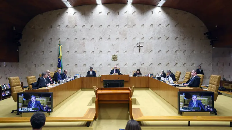 Plenário do STF durante sessão de julgamento, com ministros sentados em bancada em formato semicircular. Bandeira do Brasil, crucifixo e telas com transmissão da sessão aparecem ao fundo.