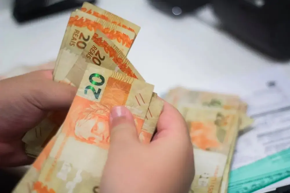 Mão segurando notas de dinheiro do tipo real, com cédulas de 20 e 10 reais e outras cédulas ao fundo em uma mesa, enquanto contas são manuseadas. Imagem usada em matéria sobre calculadora para o programa Desenrola Brasil.