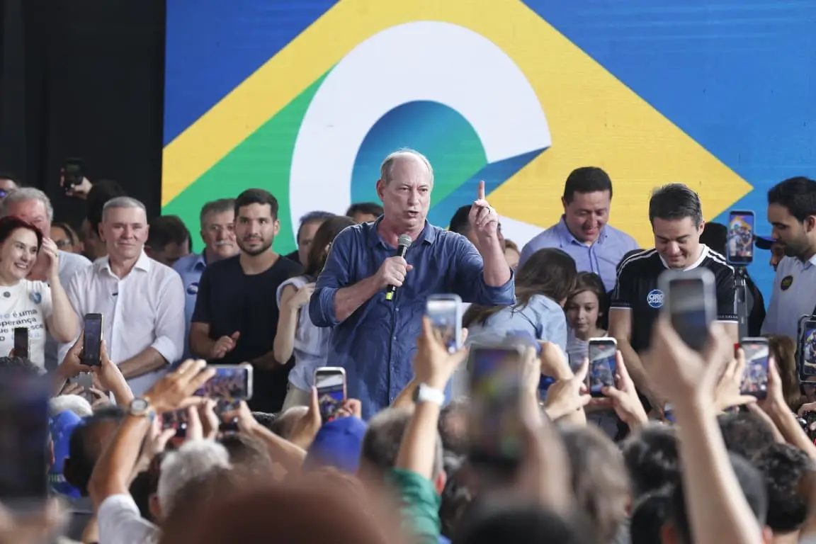 Ciro Gomes discursa em evento político diante de apoiadores, segurando um microfone e apontando o dedo para cima. Pessoas ao redor gravam o momento com celulares.