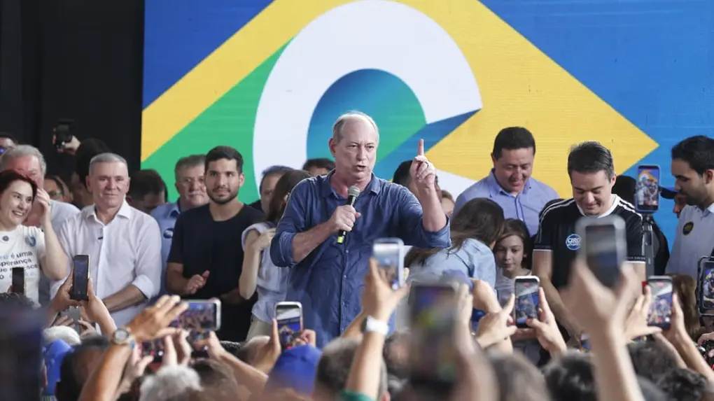Ciro Gomes discursa em evento político diante de apoiadores, segurando um microfone e apontando o dedo para cima. Pessoas ao redor gravam o momento com celulares.