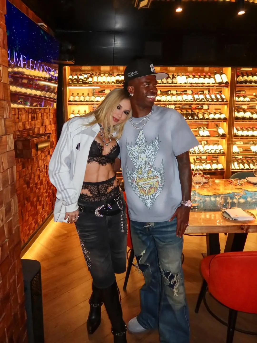 Foto posada da influenciadora Virginia Fonseca, vestindo uma blusa branca aberta e top rendado preto, ao lado do jogador Vini Jr. com blusa cinza e boné preto. Ela olha para a câmera e ele olha para o lado. Ao fundo, há uma adega iluminada com várias prateleiras de vinhos.