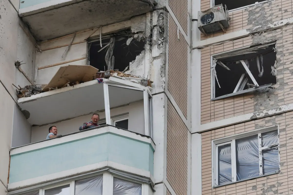 Apartamento em prédio residencial com danos estruturais visíveis: janelas quebradas, paredes destruídas e entulho exposto. Em uma varanda inferior, duas pessoas observam a área afetada enquanto parte da fachada apresenta sinais de impacto e degradação.