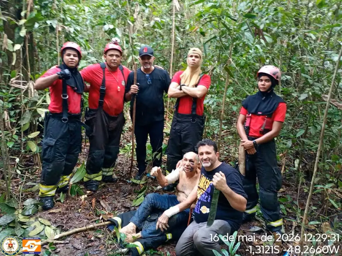 Equipe de resgate com uniformes vermelhos posa em área de mata fechada ao lado de duas pessoas sentadas no chão, em meio à vegetação densa; uma das pessoas segura um objeto, enquanto a equipe aparece equipada com capacetes e acessórios de salvamento. No canto da imagem, há data e coordenadas geográficas registradas.