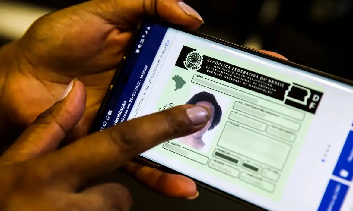 Pessoa segura um celular exibindo a versão digital da Carteira Nacional de Habilitação (CNH) na tela. A imagem mostra a interface do documento eletrônico brasileiro sendo acessada pelo aplicativo oficial.
